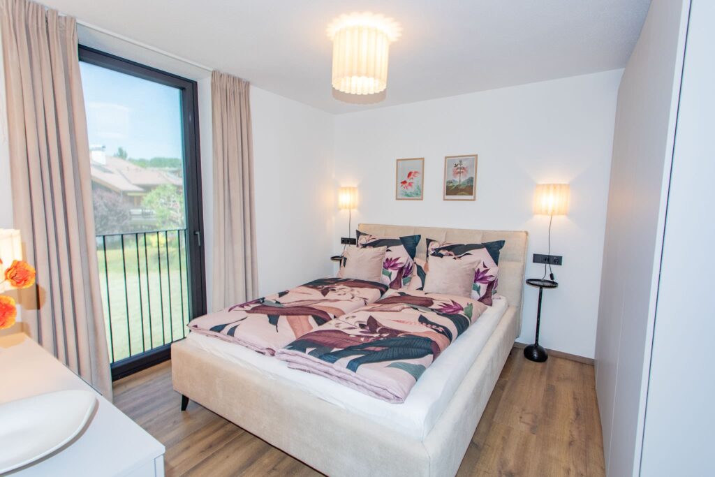 Das Bild zeigt ein Schlafzimmer mit einem Doppelbett. Auf den Betten liegen Tagesdecken mit floralen Mustern. Zwei Nachttische mit Lampen stehen seitlich vom Bett. Ein Fenster mit Geländer bietet einen Ausblick auf eine Landschaft. Zwei Gemälde mit floralen Motiven hängen an den Nachttischen. Der Boden ist aus Holz.