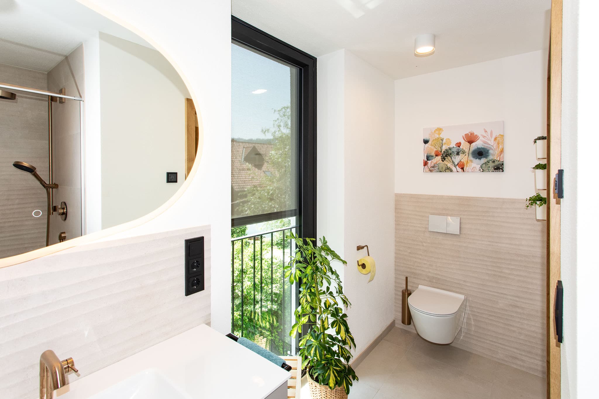 Das Bild zeigt einen Teil eines Badezimmers. Im Zentrum befindet sich ein Waschtisch mit einem Spiegel, Armatur und Handtuchhalter. Ein schwarzer Fensterrahmen ist rechts im Bild zu erkennen, der einen Ausblick auf eine Gartenfront bietet. Im Vordergrund sind Pflanzen und ein Körbchen zu sehen. Die Beleuchtung ist durch einen Lichtkreis im Spiegel erzeugt.
