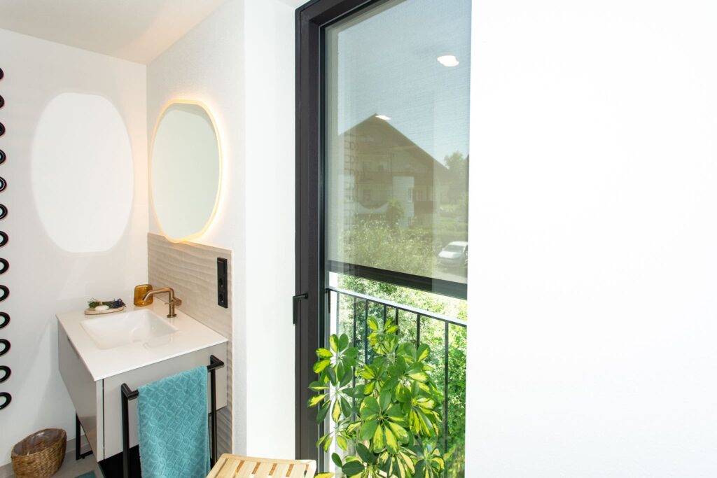 Das Bild zeigt einen Teil eines Badezimmers. Im Zentrum befindet sich ein Waschtisch mit einem Spiegel, Armatur und Handtuchhalter. Ein schwarzer Fensterrahmen ist rechts im Bild zu erkennen, der einen Ausblick auf eine Gartenfront bietet. Im Vordergrund sind Pflanzen und ein Körbchen zu sehen. Die Beleuchtung ist durch einen Lichtkreis im Spiegel erzeugt.