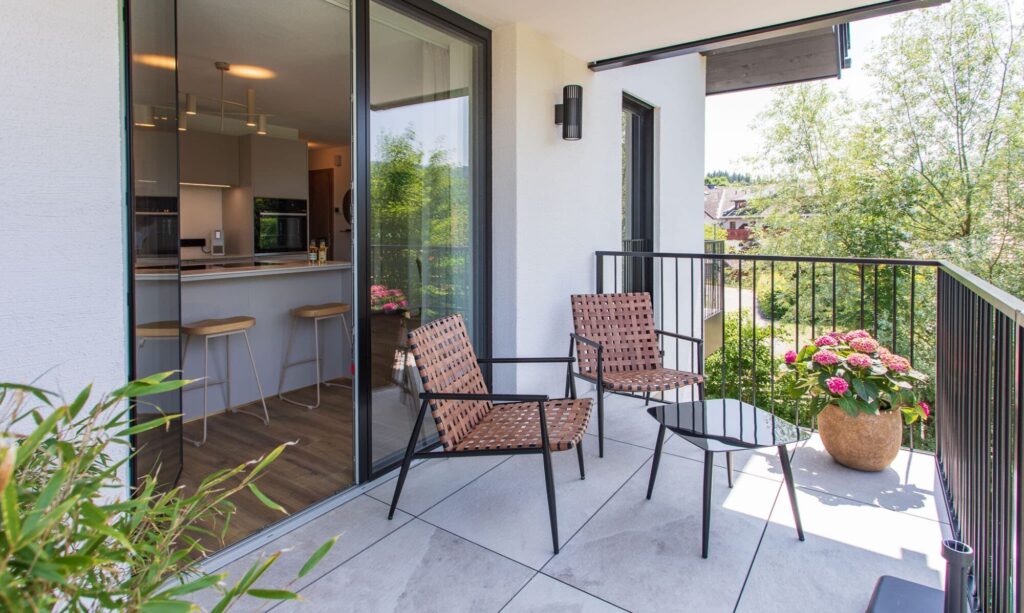 Balkon mit Blick auf eine moderne Küche. Auf dem Balkon stehen zwei Sitzstühle und ein Tisch. Im Hintergrund sind die Küche mit einem Kühlschrank und einer Arbeitsplatte sowie ein Außenbereich mit Fenstern zu sehen. Der Balkon ist mit weißem Fliesenbelag bedeckt und von Pflanzen umgeben